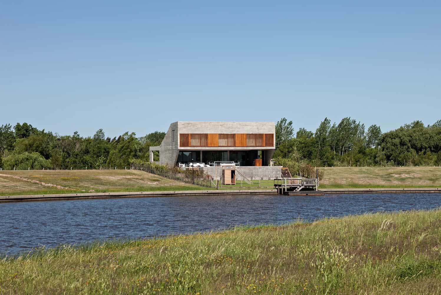 Contemporary waterfront house with modern architectural design, concrete structure, large glass windows, and wooden accents, situated by a serene river with lush greenery.
