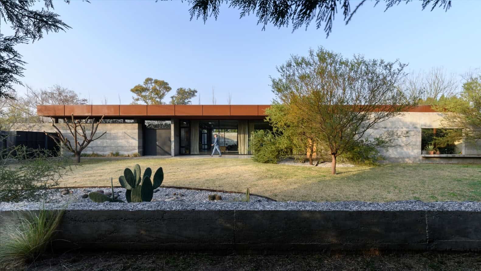 Modern desert-style house with minimalist architecture, concrete walls, flat roof, and large glass windows, surrounded by desert plants and cacti, showcasing contemporary architectural design.