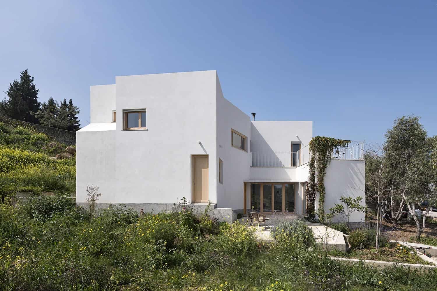 Modern minimalist white house with geometric architectural design and large windows, surrounded by lush greenery and garden landscape.