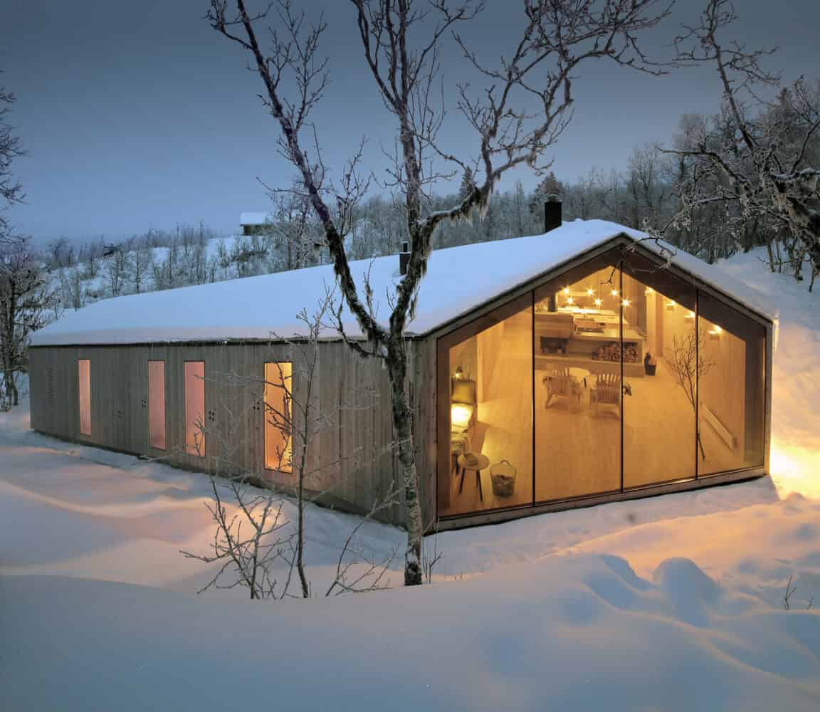 Cozy wooden house with large glass windows illuminated inside during winter snowfall, set in a snowy landscape with leafless trees, showcasing warm modern architecture and inviting interior design.