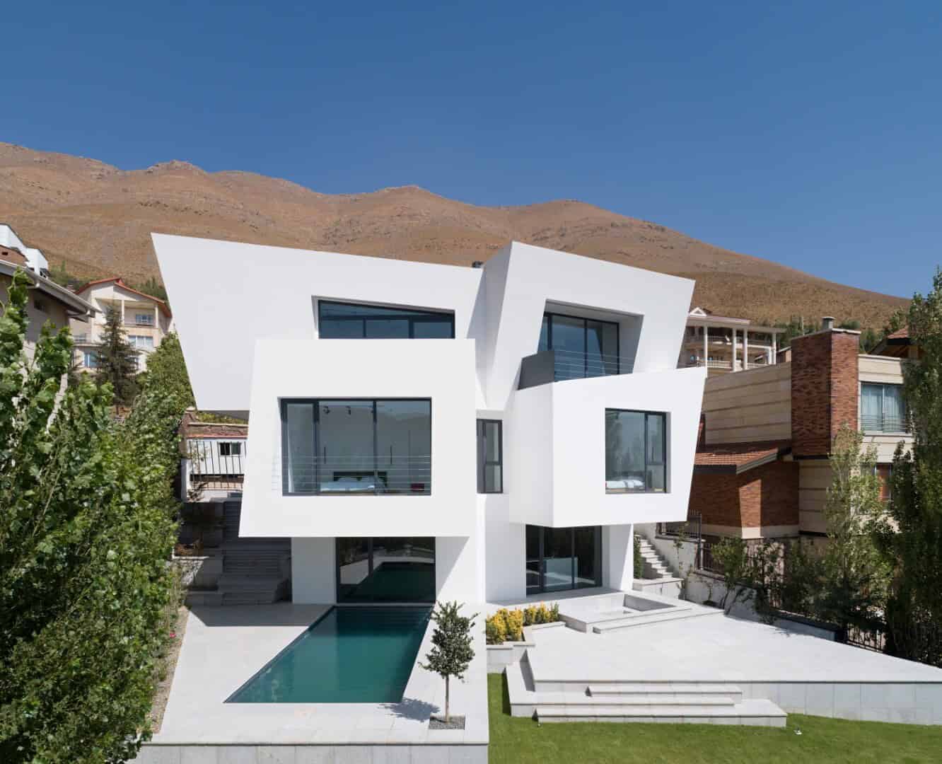 Contemporary modern white residential house with geometric design and large glass windows, featuring an outdoor swimming pool, situated in a hillside neighborhood with scenic mountain views.