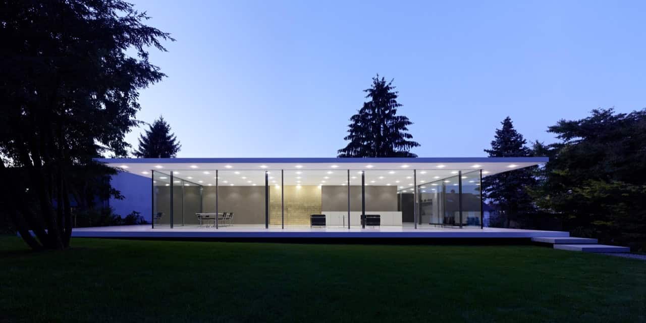 Sleek modern glass house with open interior design and minimalist architecture, surrounded by lush trees and a well-maintained lawn, ideal for contemporary architecture and luxury home design.