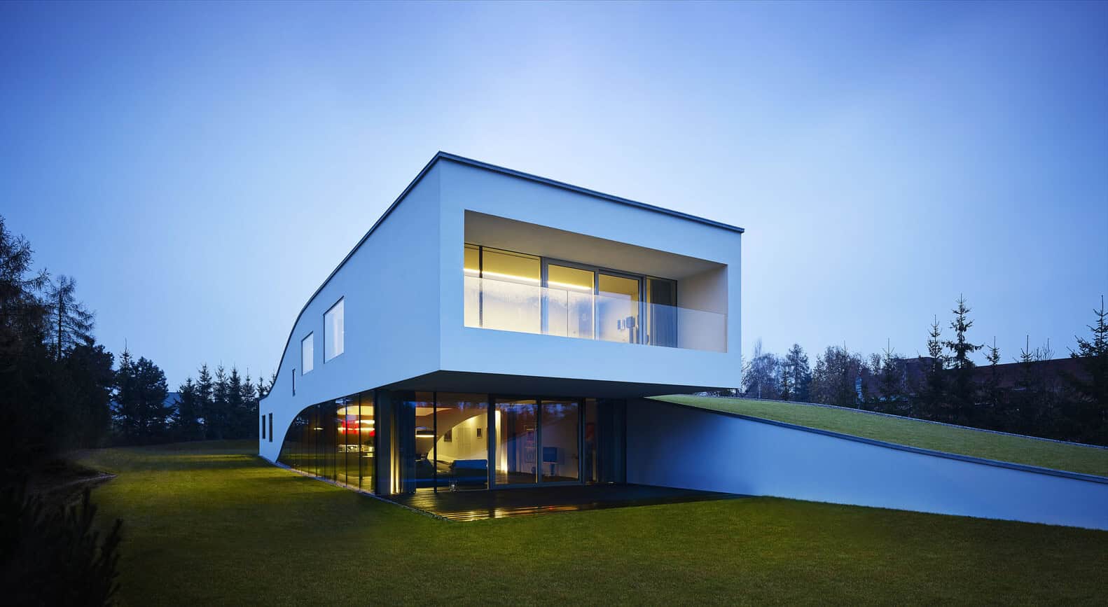 Modern minimalist white house with geometric design and large glass windows, surrounded by green lawn and trees at dusk.