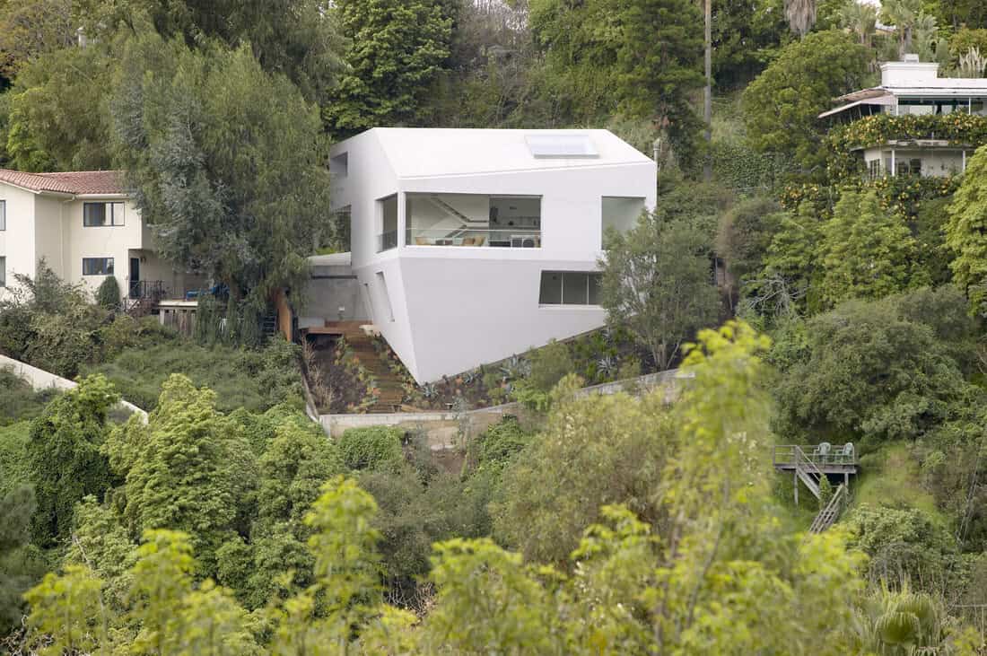 Modern white hillside house with angular architectural design nestled among lush greenery in a scenic residential area.