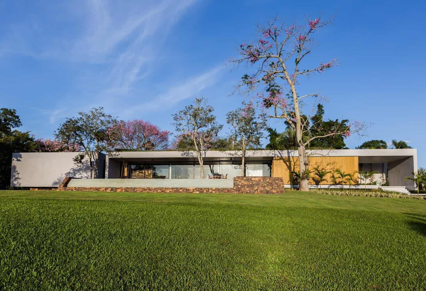 Modern minimalist house with clean lines and large glass windows in a lush green landscape under a clear blue sky.