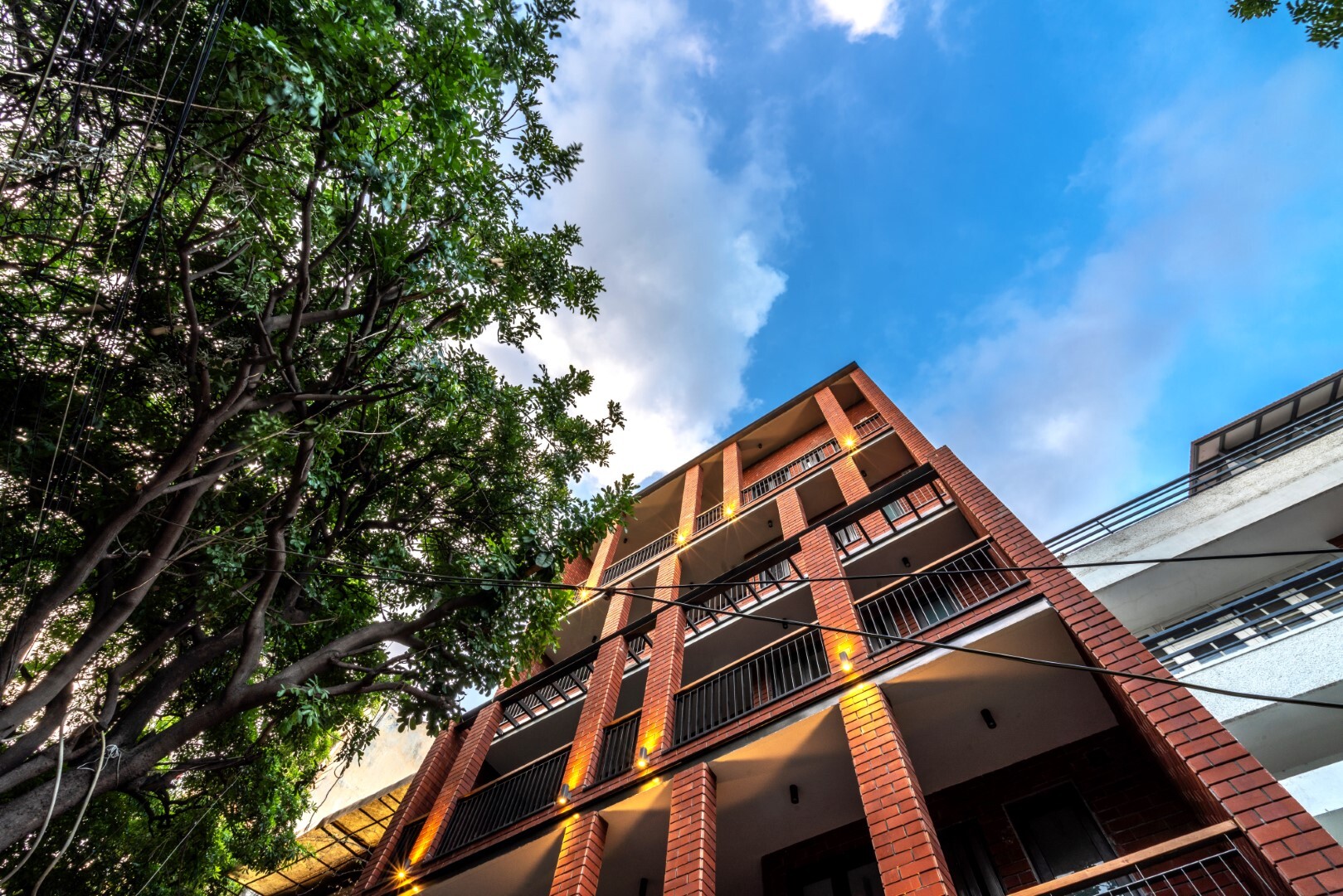 Elegant modern multi-story building with brick facade and balconies, set against a vibrant blue sky with lush green trees, showcasing contemporary architecture and urban design.