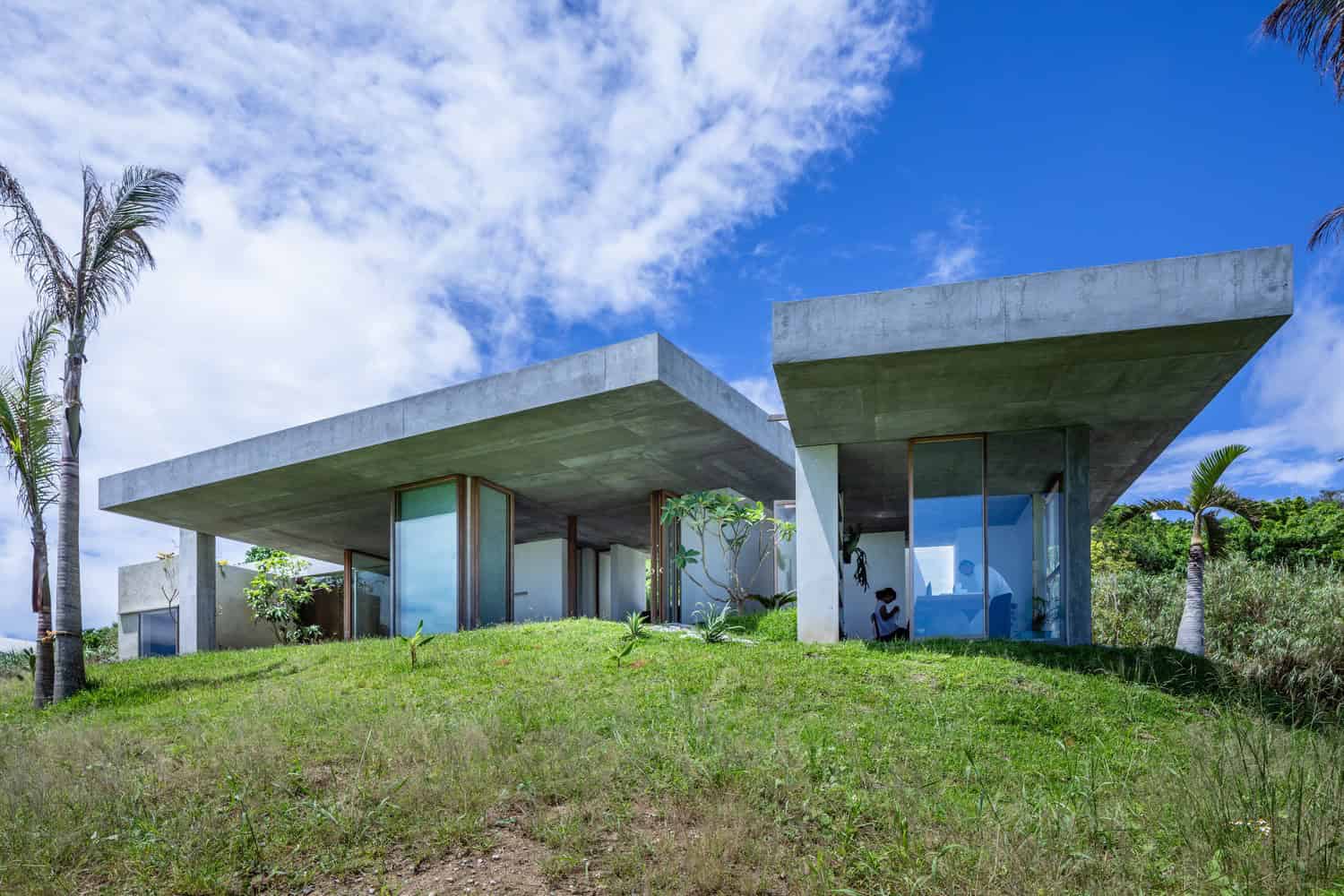 Contemporary modern house with concrete and glass architecture, featuring minimalist design, clean lines, and open spaces set on a lush green hillside under a bright blue sky.