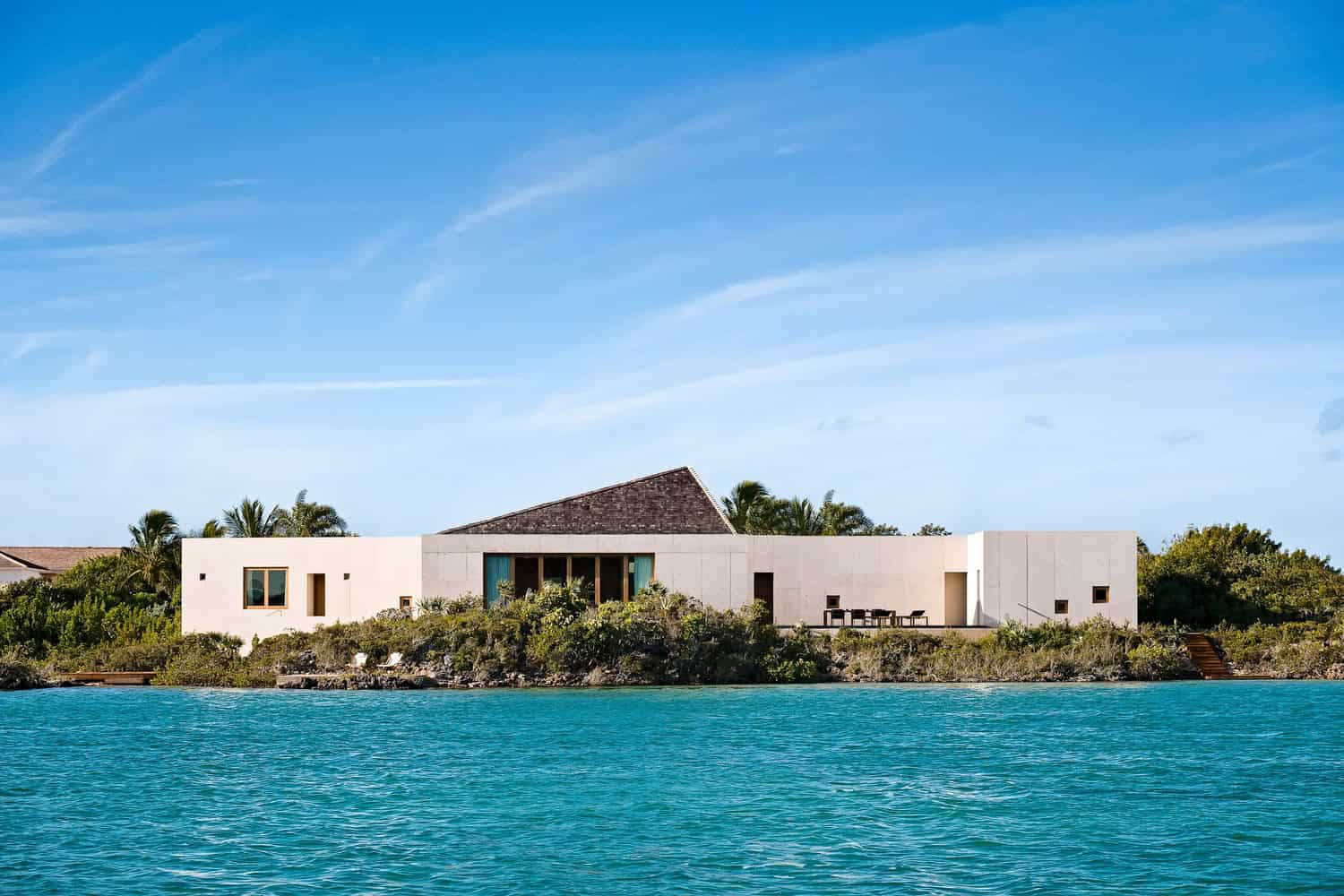 Stunning modern waterfront house with minimalist design, large glass windows, and outdoor terrace surrounded by lush greenery and tropical landscape, overlooking clear blue ocean waters.