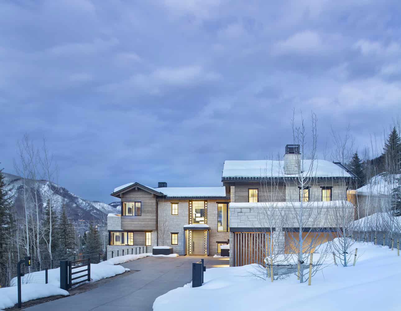 Contemporary mountain home with modern architecture, wooden and stone exterior, large windows, snow-covered landscape, and scenic backdrop of snow-capped mountains and trees.