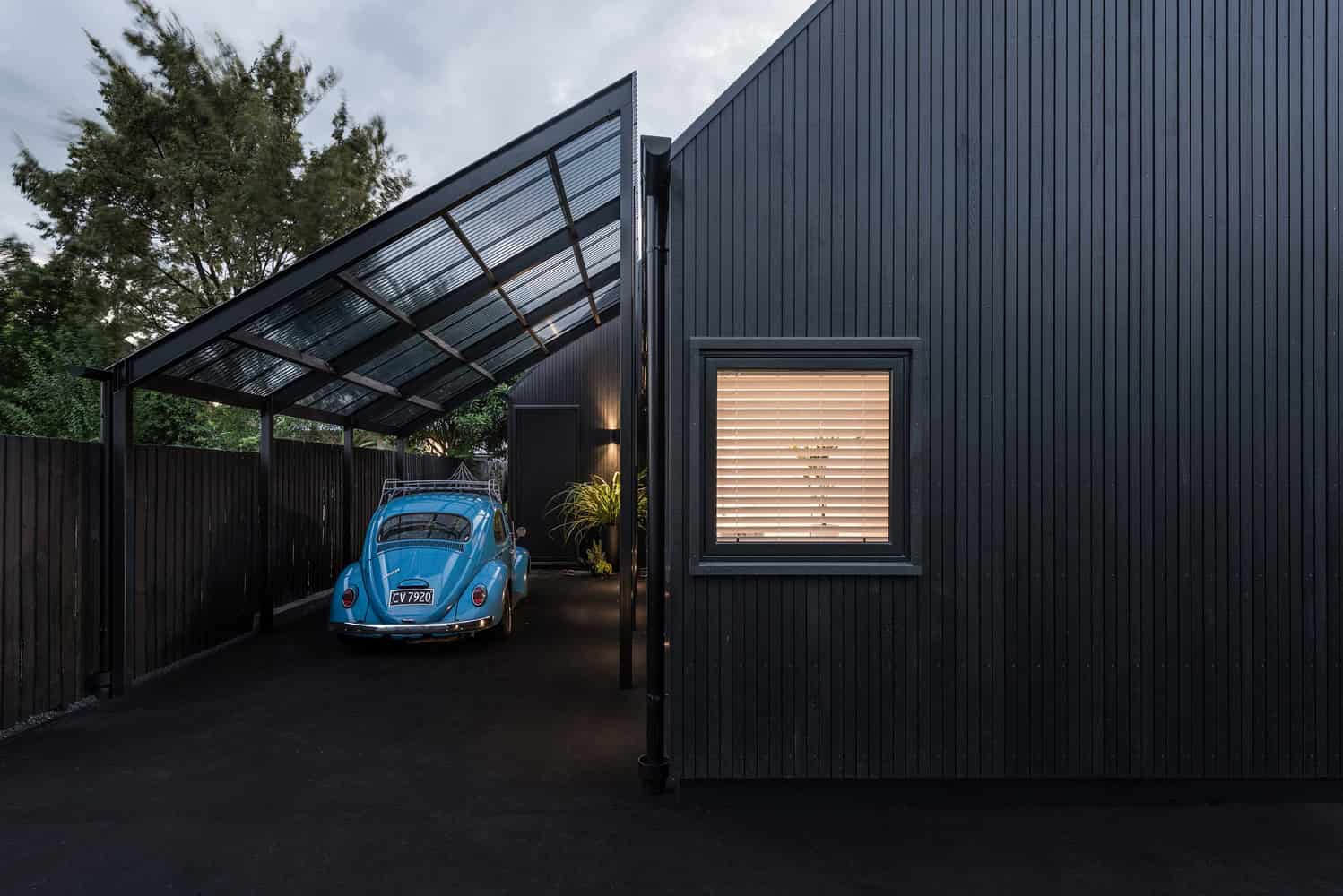 Modern black house with sleek vertical siding and a stylish carport featuring a retro blue car, outdoor lighting, and window with blinds, exemplifying contemporary architecture and innovative design.