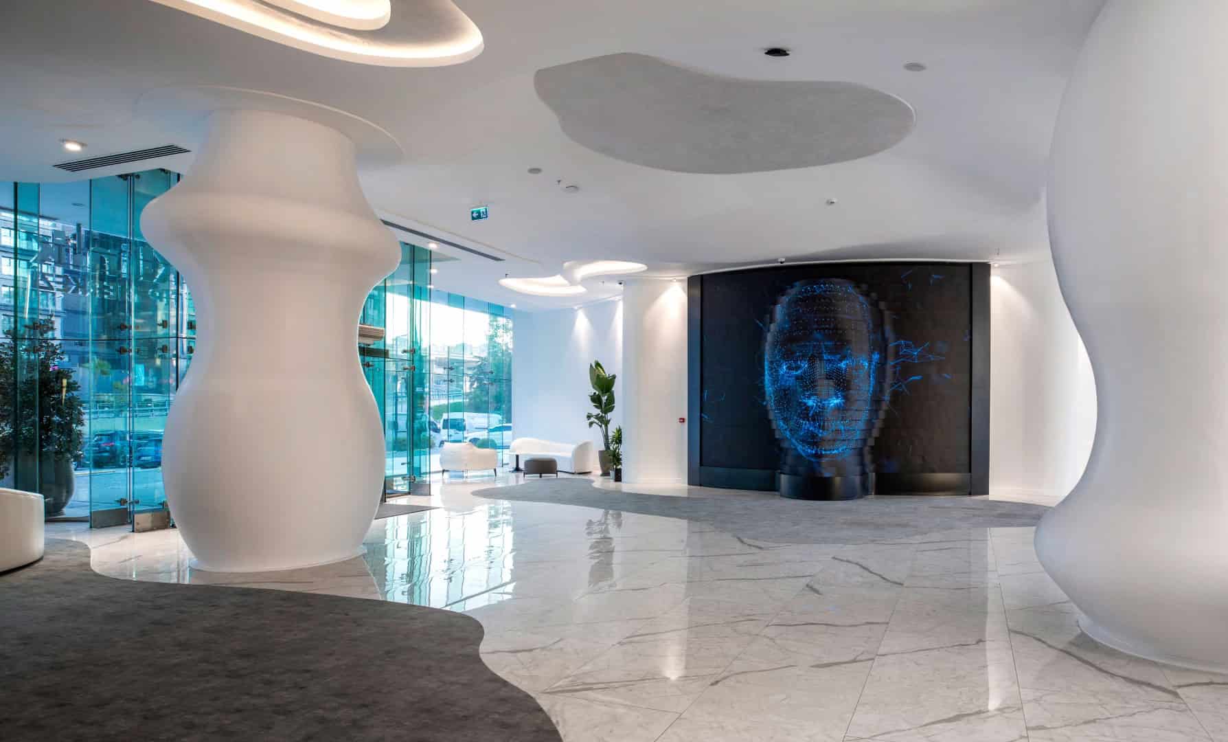 Futuristic hotel lobby with modern white sculptural pillars, sleek marble flooring, and a striking digital face sculpture illuminated in blue, showcasing contemporary architecture and innovative interior design.