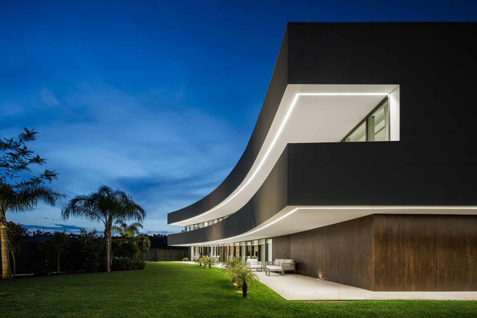 Contemporary modern house with sleek architectural design, curved white exterior roof and extensive outdoor patio at dusk.