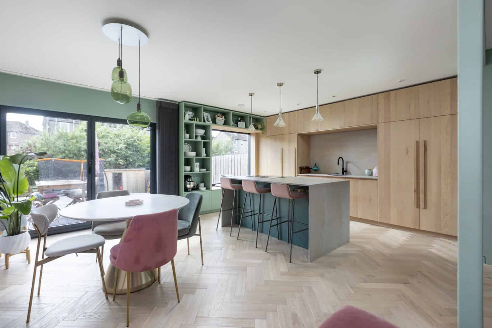 Modern open-concept kitchen and dining area with pastel pink and gray chairs, wooden cabinetry, and a stylish kitchen island featuring sleek pendant lighting and contemporary design elements.
