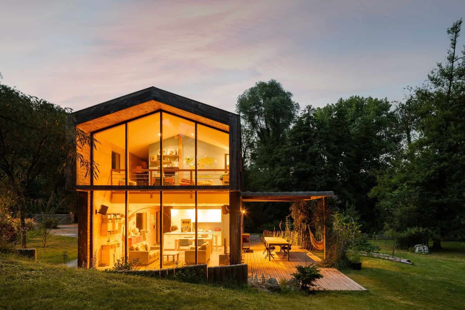 Contemporary eco-friendly house with large glass windows and modern interior design set in lush greenery at dusk.