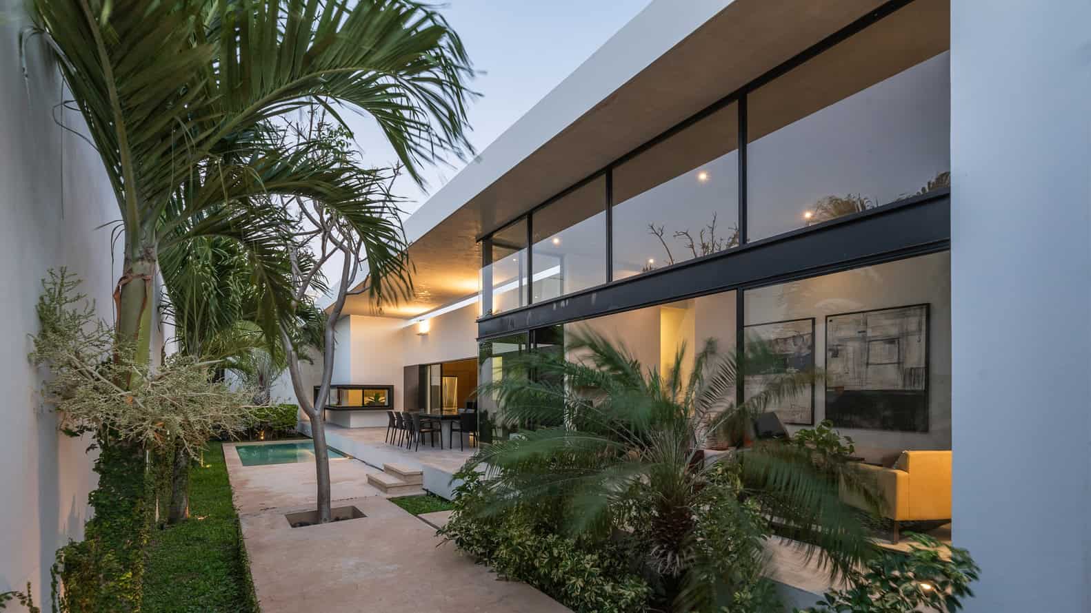 Contemporary modern house exterior with sleek glass walls, lush tropical landscaping, and cozy outdoor seating, highlighting innovative architecture and stylish design elements.