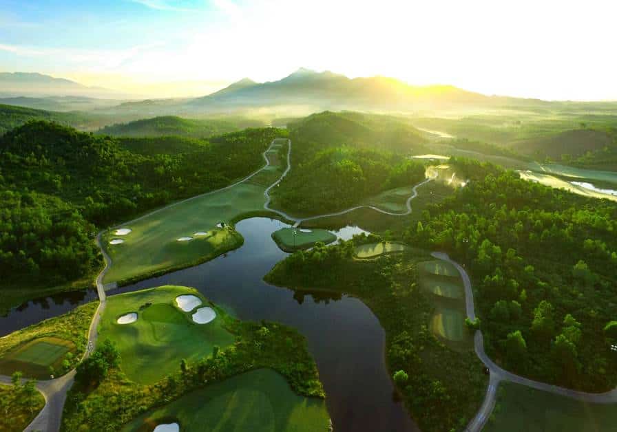 Aerial view of a golf course with lush green fairways, water hazards, and scenic mountain backdrop during sunrise, showcasing beautiful landscape architecture and outdoor design elements.