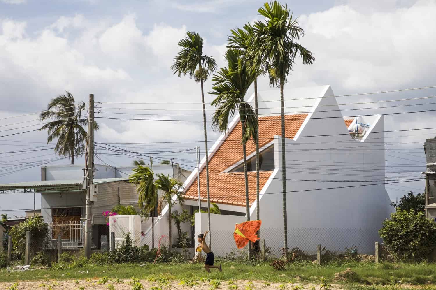 Modern architectural house with unique angular design and orange tiled roof, set against cloudy sky, surrounded by palm trees and vibrant greenery, showcasing innovative residential architecture.