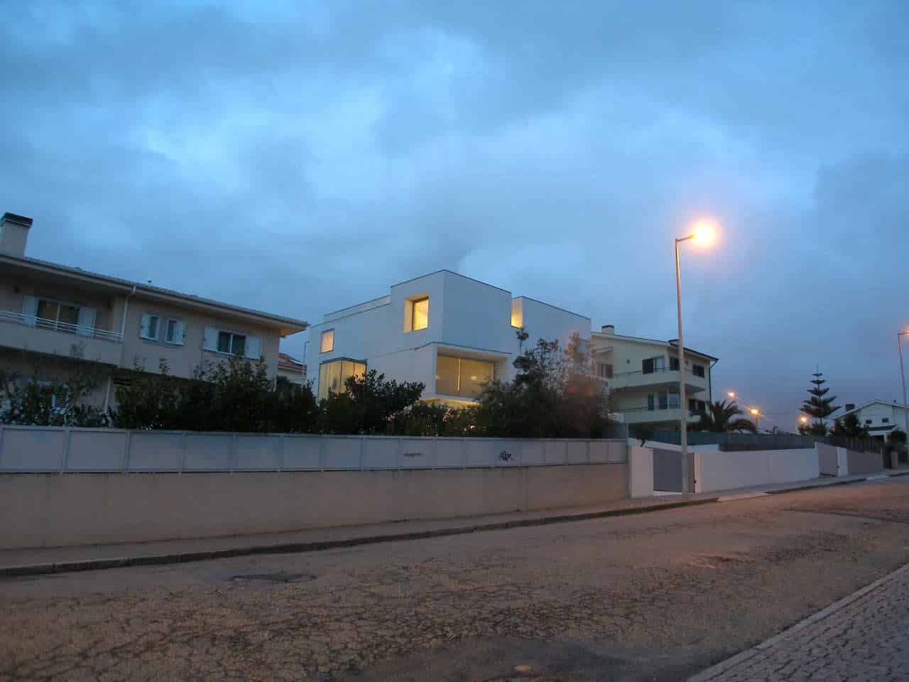 Contemporary modern white house architecture with minimalist design and large glass windows, illuminated at dusk, surrounded by a white fence on a paved street.