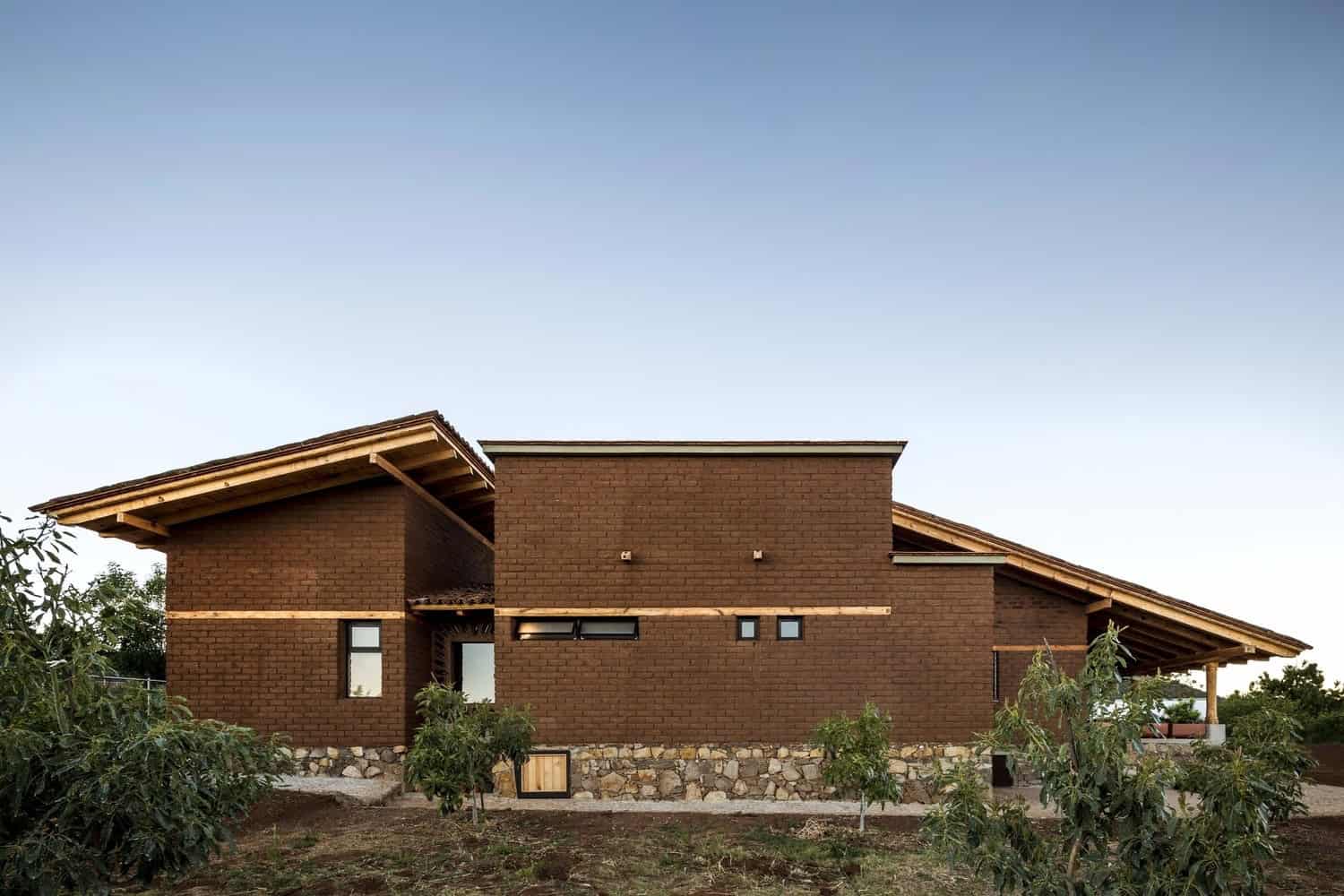 Modern brick house with sloped wooden roof, stone foundation, and large windows, surrounded by greenery and under a clear sky.