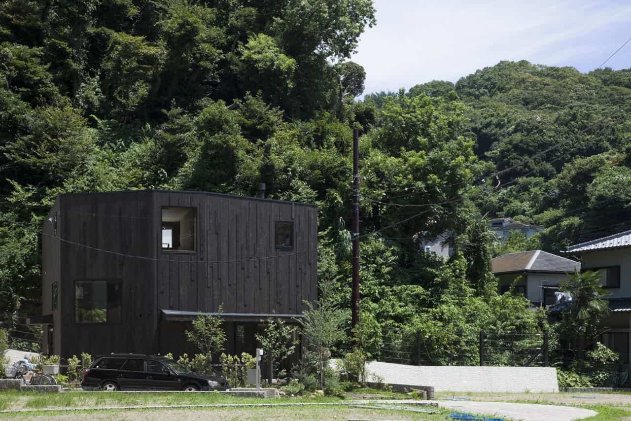 Modern black wooden residential house with unique geometric design, surrounded by lush green trees in a hilly landscape.