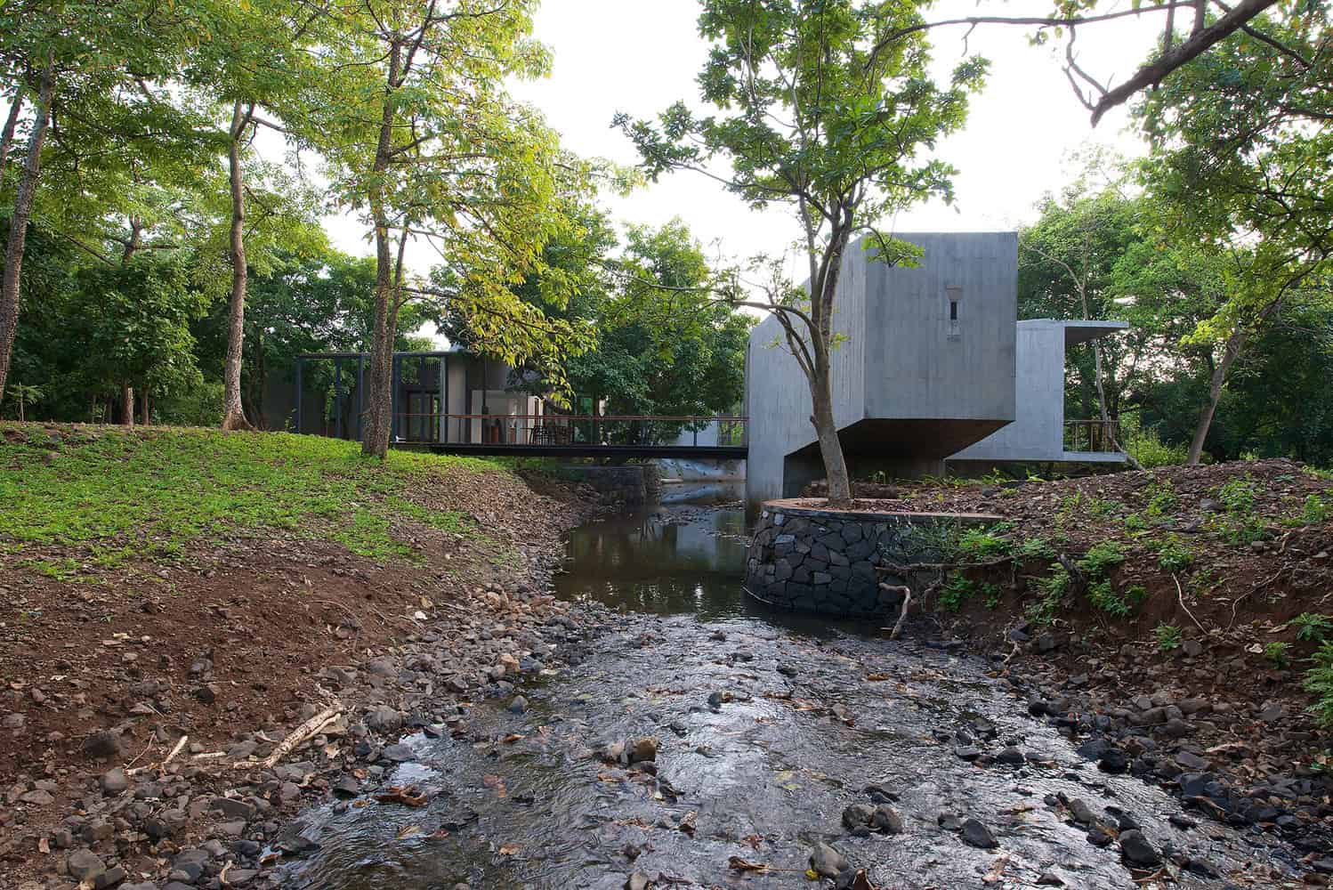Modern concrete house with sleek design nestled in lush green forest, featuring large windows, minimalist architecture, and a serene natural creek setting for eco-friendly living.