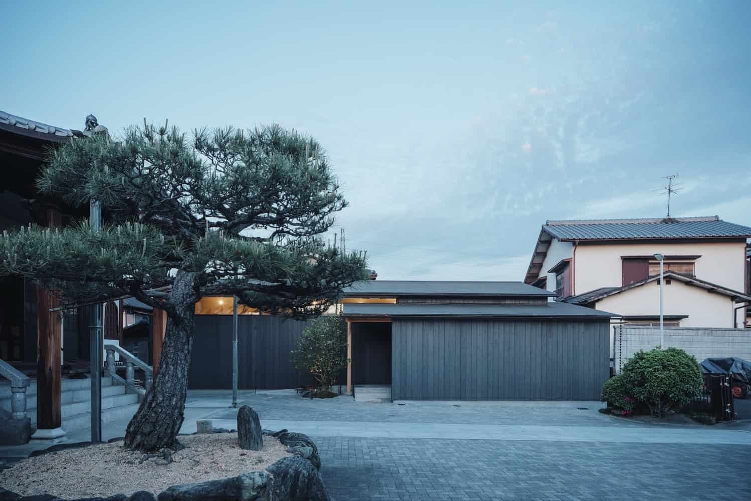 Rustic modern house exterior with traditional Japanese landscaping and minimalist architecture, featuring a pine tree, stone pathway, and clean architectural lines.