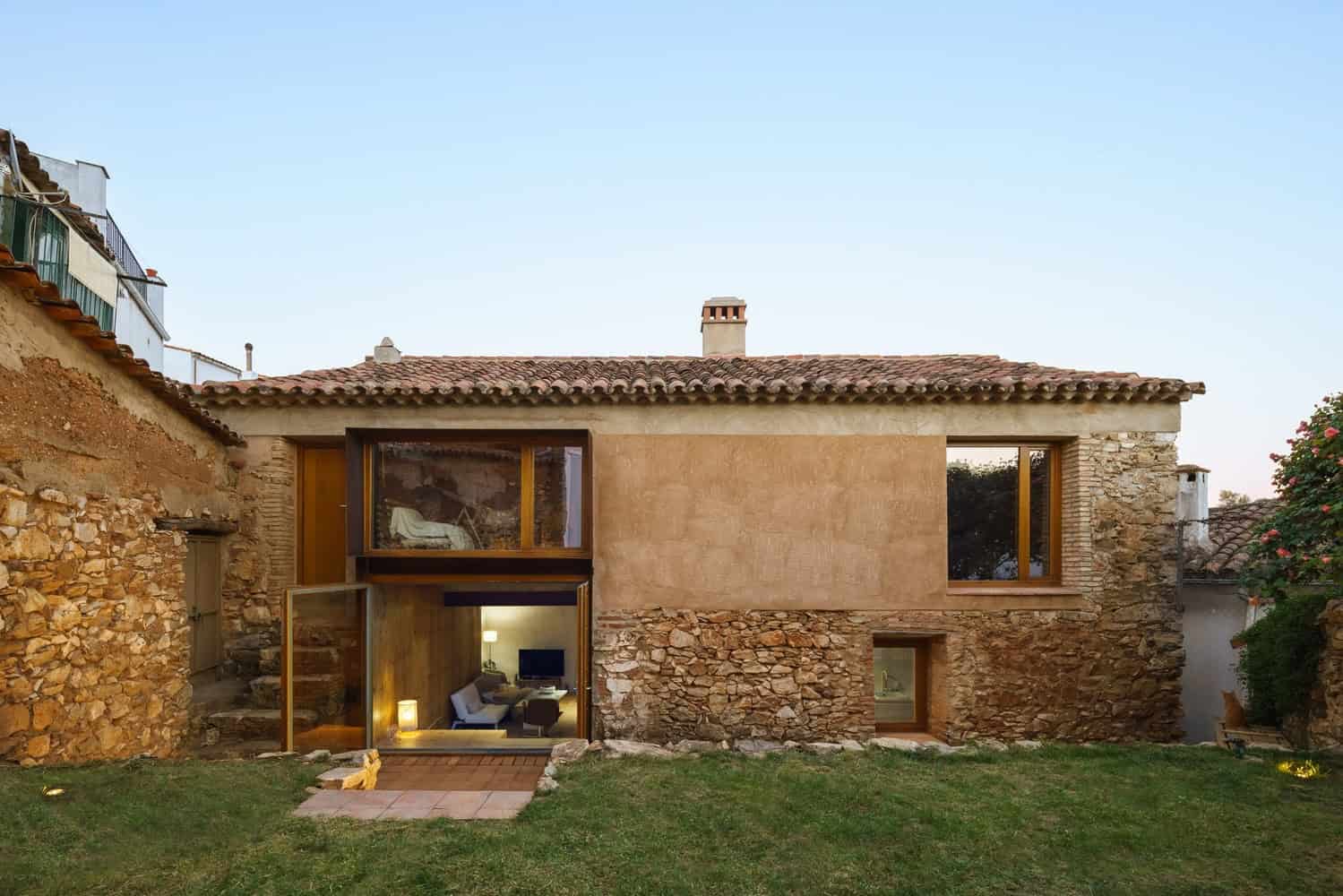 Rustic modern house with stone exterior, large wooden framed windows, and a cozy interior visible from the garden. Traditional tile roof complements contemporary design elements.