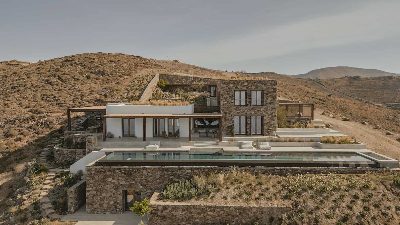 Modern desert hillside luxury home with infinity pool, stone and white exterior, expansive windows, and outdoor lounging area, blending contemporary architecture with natural landscape.