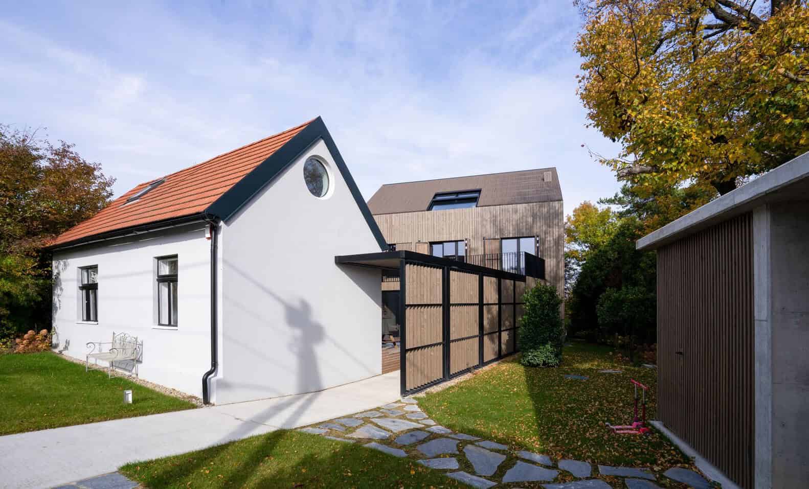 Contemporary residential architecture featuring a white gable house with a red tile roof, wooden modern extension, and landscaped yard with fall foliage.