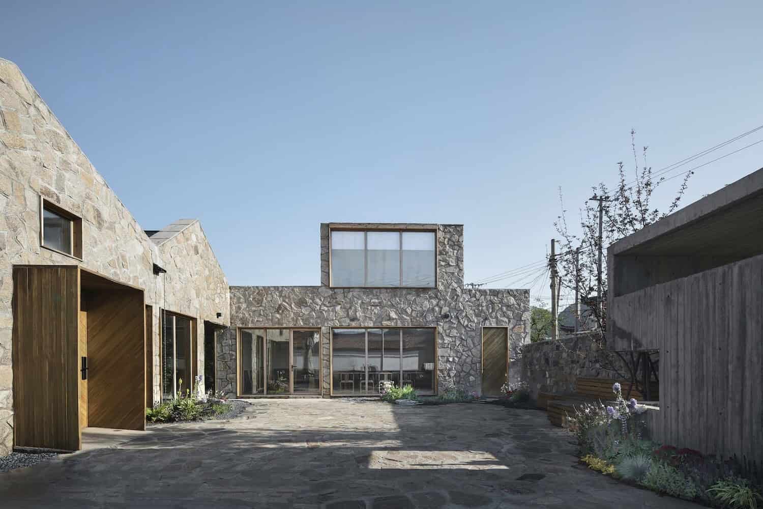 Stone modern house with large glass windows and unique architectural design, featuring a combination of natural stone and wood accents, set in a minimalist outdoor courtyard with plants and benches.