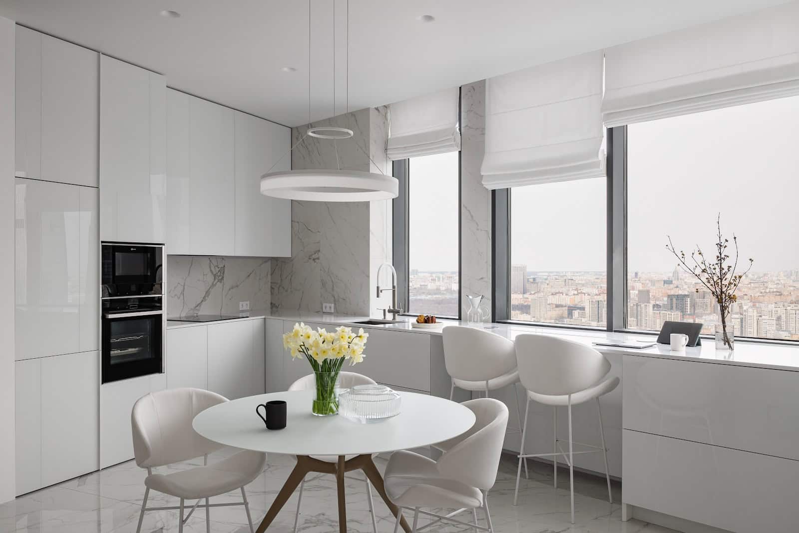 Modern minimalist white kitchen with large windows, marble accents, and contemporary furniture, showcasing luxury interior design and urban cityscape views.