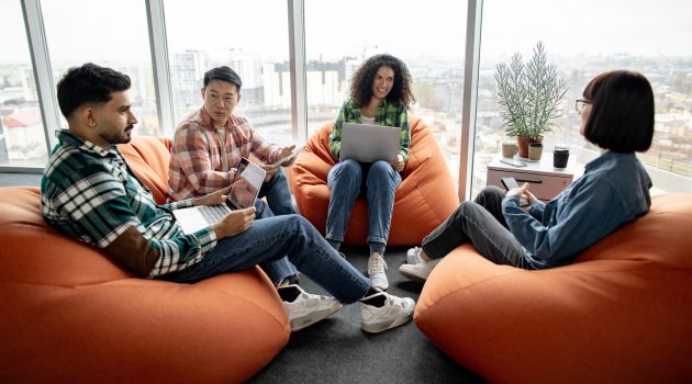 Diverse team of managers in casual wear having conversation while resting on cozy poufs in creative office. Confident collaborators improving corporate strategy using modern technologies in workplace.