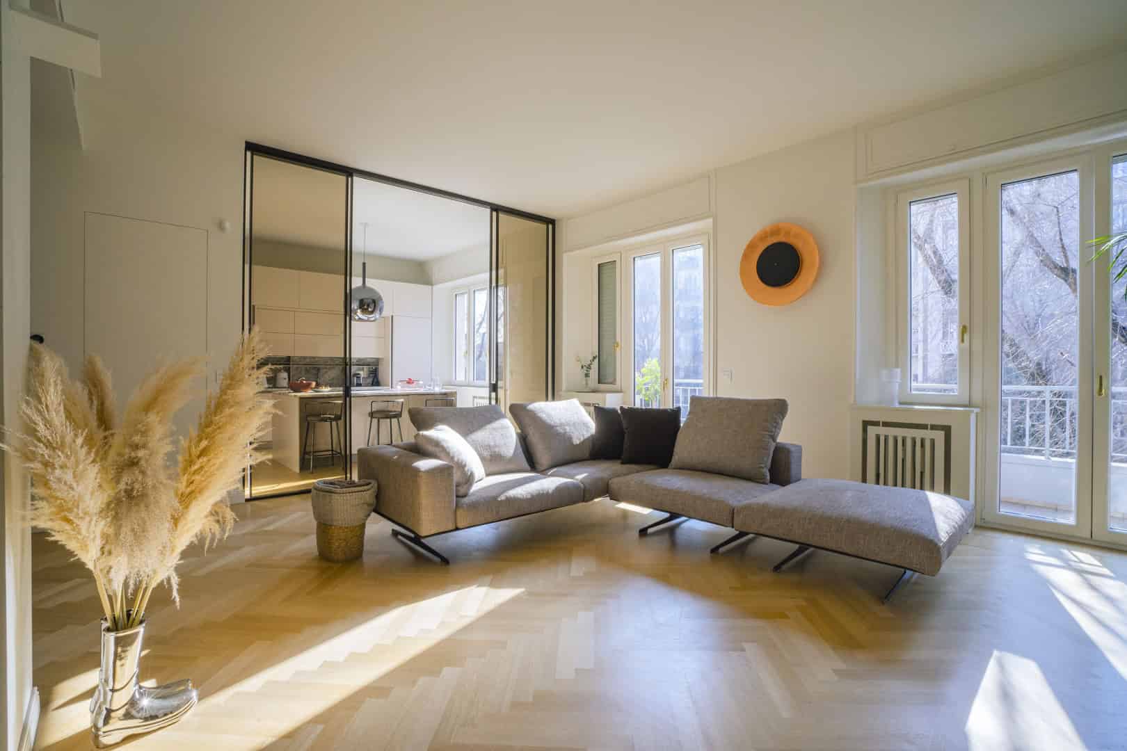 Modern open-concept living room with neutral-toned furniture and large windows letting in natural light, featuring a minimalist aesthetic and seamless flow to the kitchen area.