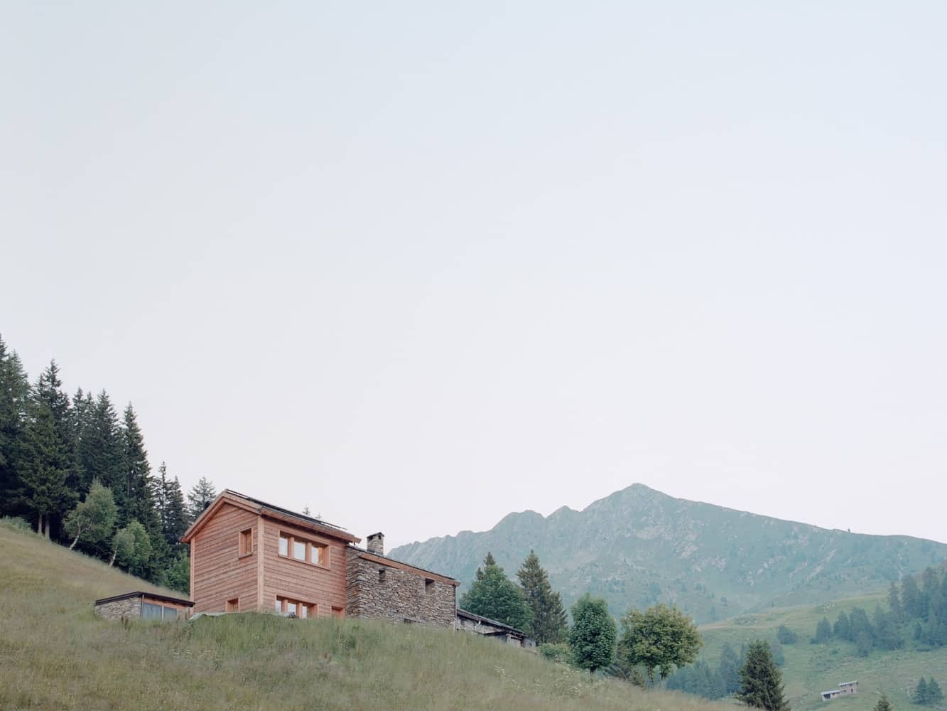 Rustic mountain cabin with modern wooden accents nestled on a lush green hillside, surrounded by tall trees and scenic mountain views, exemplifying harmony between architecture and nature.