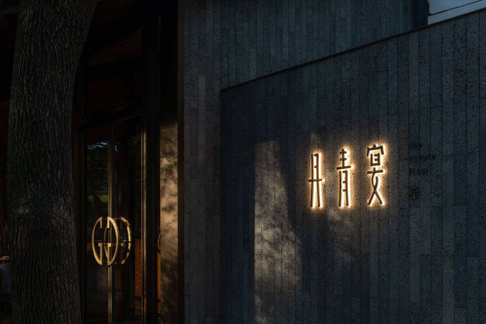 Contemporary building entrance with illuminated Asian-inspired characters and modern architectural design, featuring textured concrete walls and sleek glass doors.