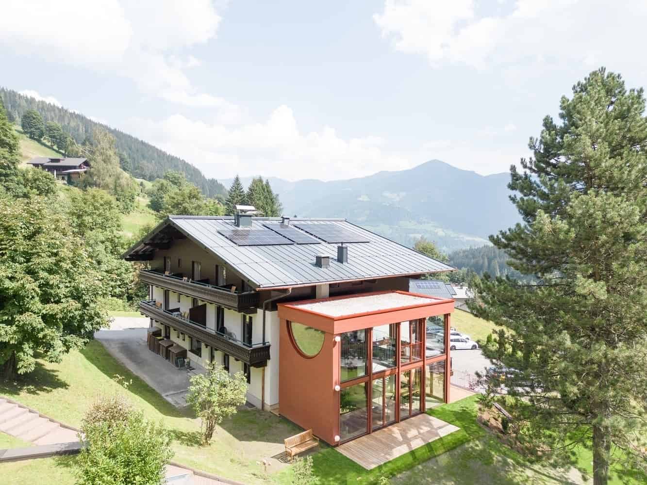 Modern alpine hotel with solar panels, surrounded by lush greenery and mountain views, showcasing contemporary architecture blending with natural landscape.