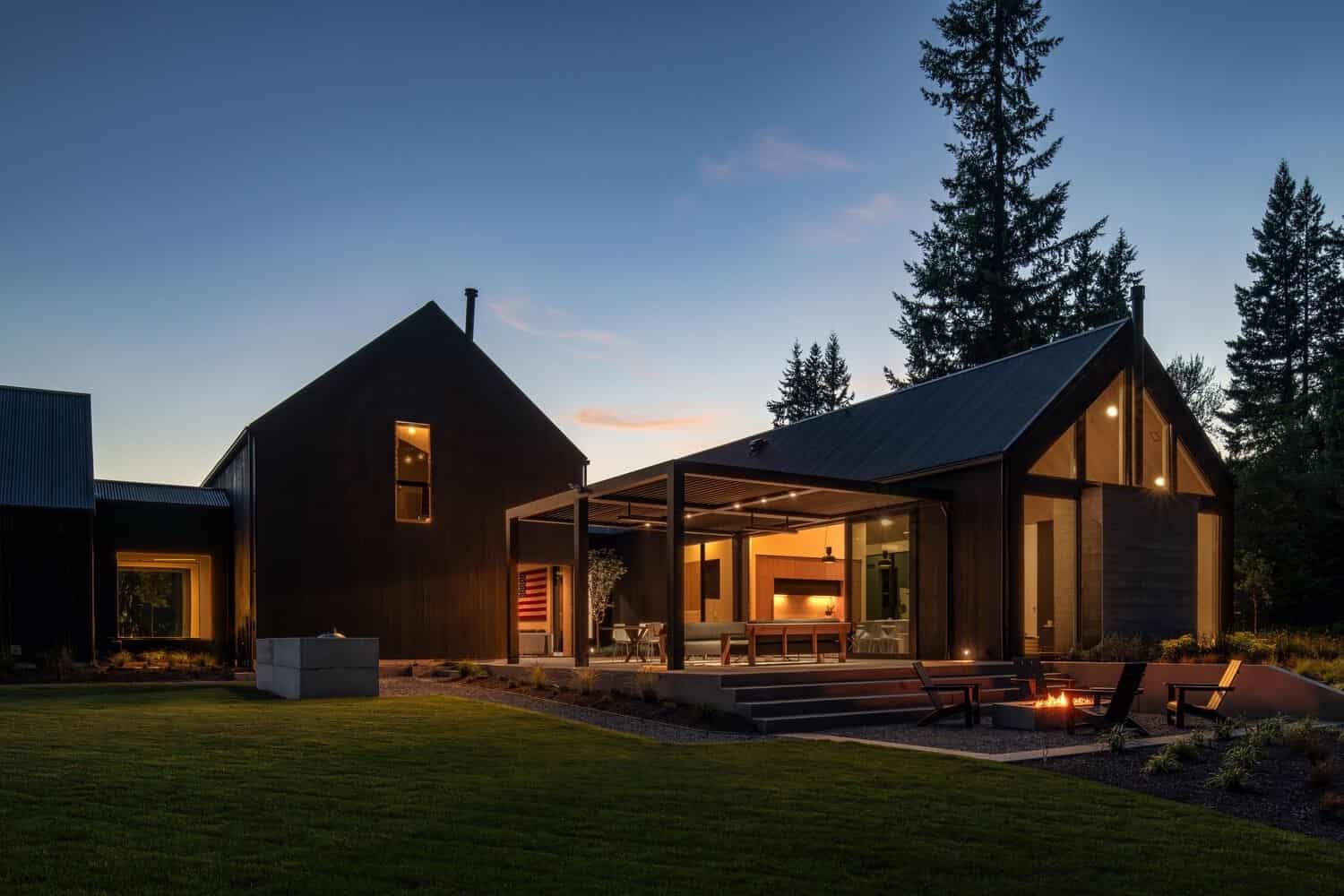 Modern black farmhouse with outdoor living space and fire pit at dusk, surrounded by trees and lush green lawn, showcasing contemporary architectural design and stylish landscaping, perfect for luxury home enthusiasts.