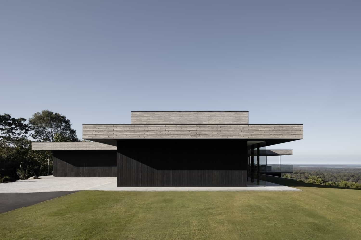 Modern minimalist house with black and gray concrete exterior, flat roof, and expansive glass windows, set against a clear blue sky and lush green lawn.
