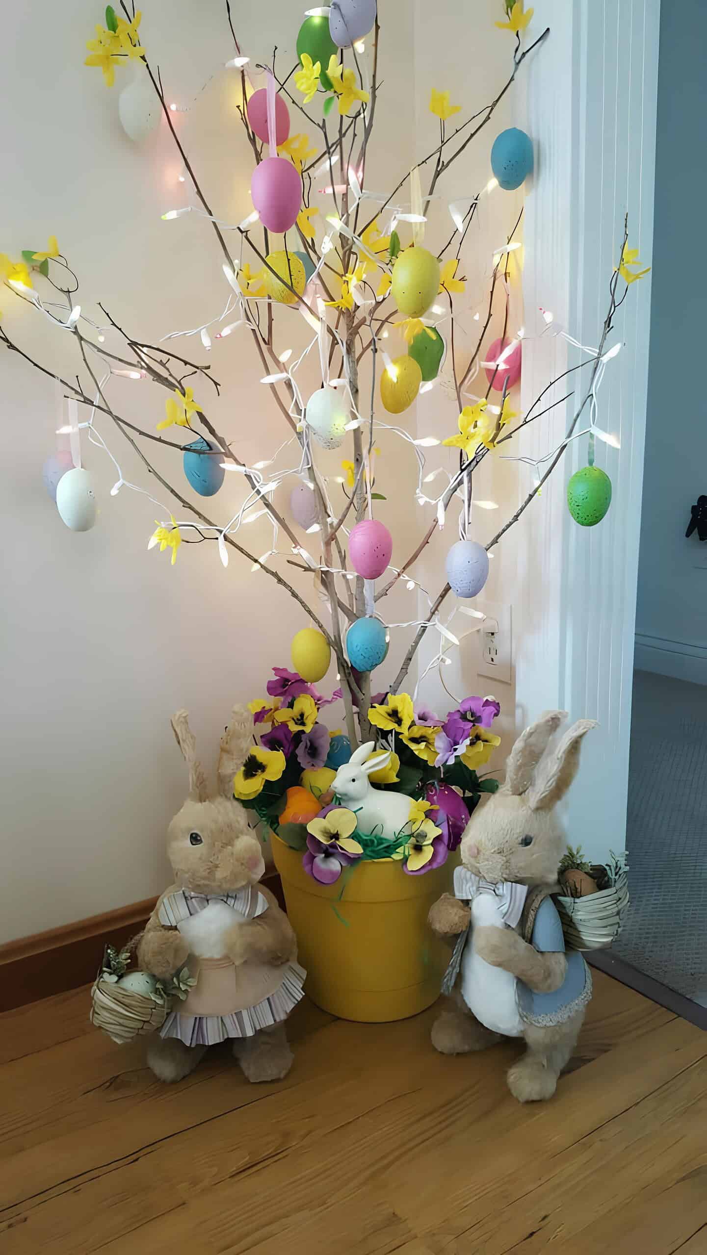 6. Creative Easter Tree DIY Easter egg tree made with branches, pastel hanging eggs, yellow flower accents, and string lights in a yellow pot, surrounded by plush bunny figurines and artificial spring flowers – festive indoor Easter decoration idea.