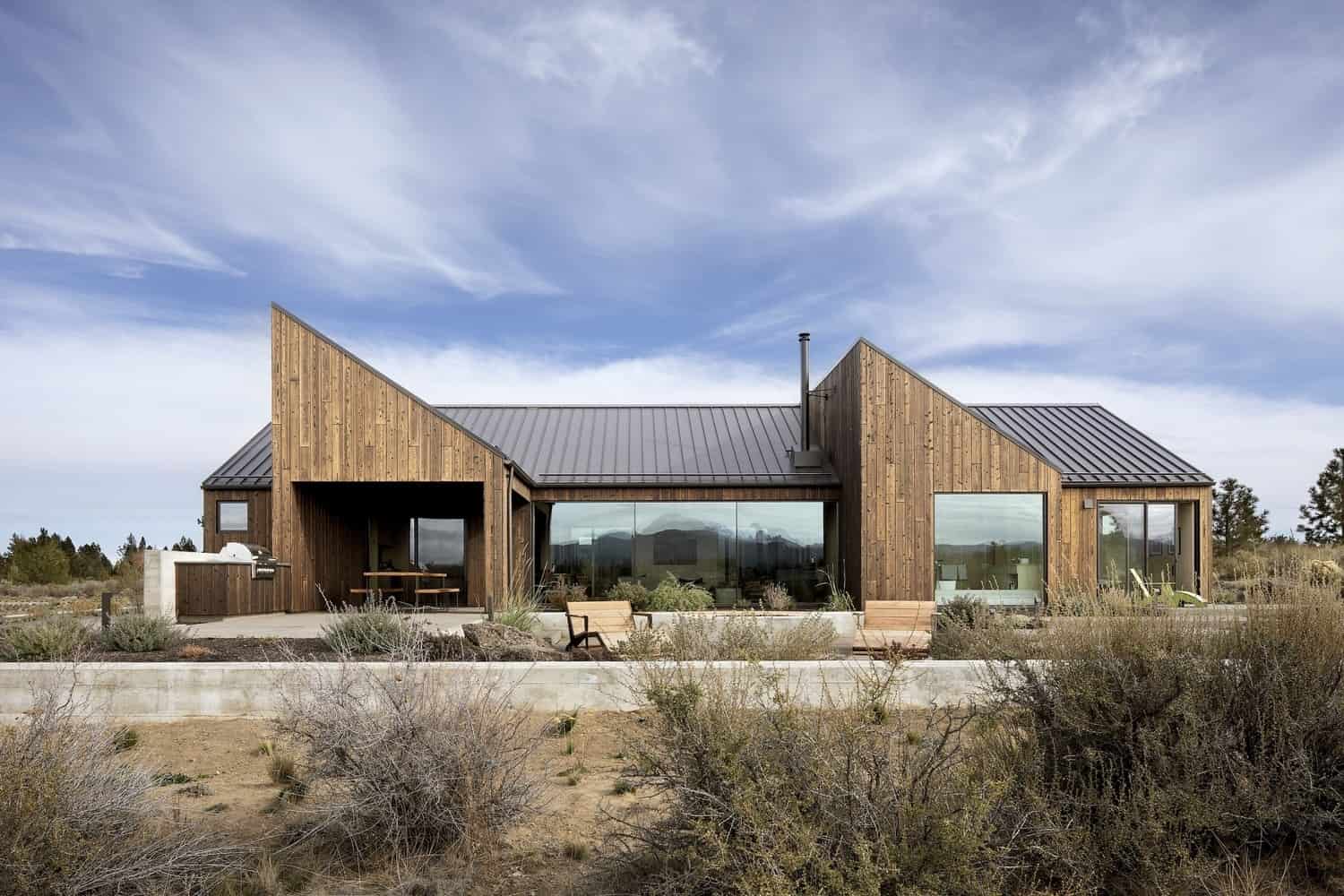 Modern rustic house with wooden siding, large glass windows, and metal roof, surrounded by desert landscape, showcasing contemporary architectural design and outdoor living space.