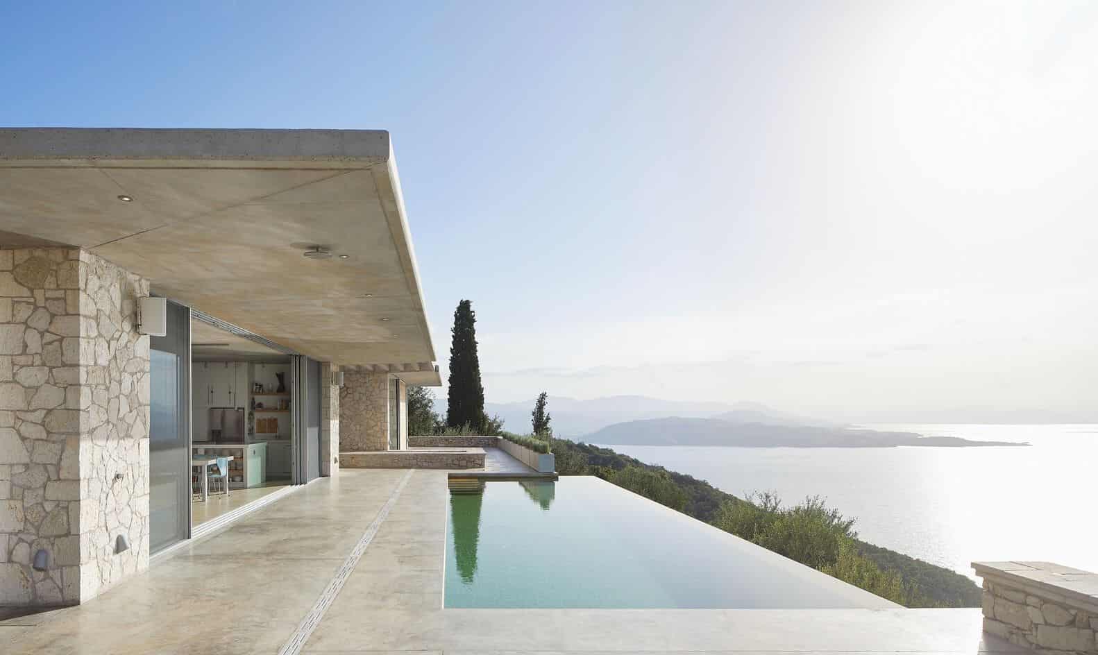 Modern luxury house with infinity pool overlooking scenic landscape and sea view, featuring contemporary architecture, stone wall accents, and floor-to-ceiling glass doors.