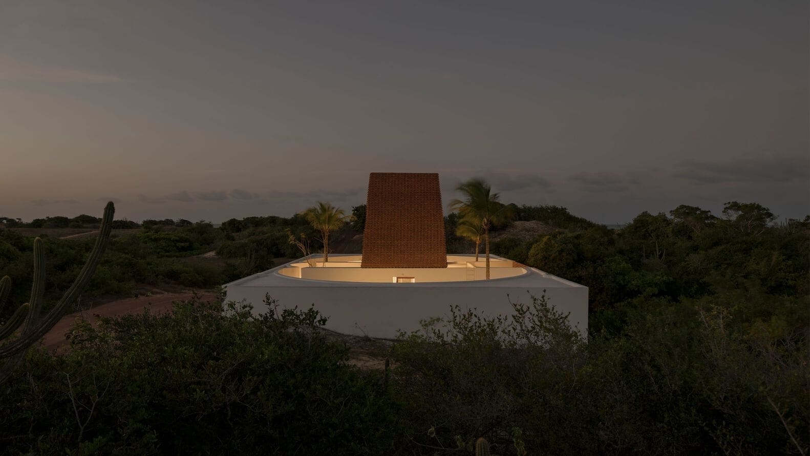 Modern architectural house with a unique rounded white structure, surrounded by desert vegetation and palm trees, featuring a tall brick chimney-like element at sunset.