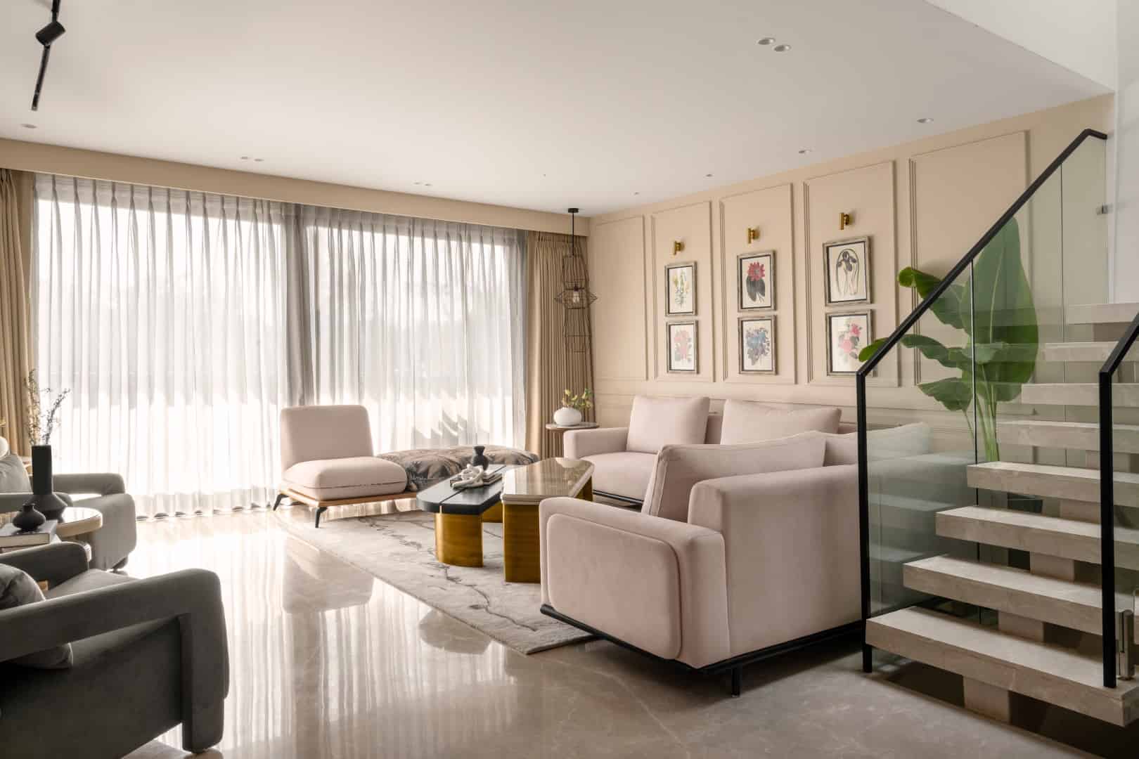 Bright modern living room with beige and grey furniture, large windows with sheer curtains, stylish wall art, and elegant staircase with glass railing, showcasing contemporary interior design and cozy aesthetic.