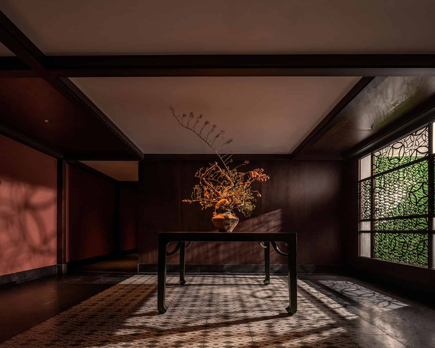 Elegant interior with a minimalist black table and an artistic flower arrangement, featuring intricate window patterns casting shadows on the tiled floor, showcasing modern Asian-inspired design.
