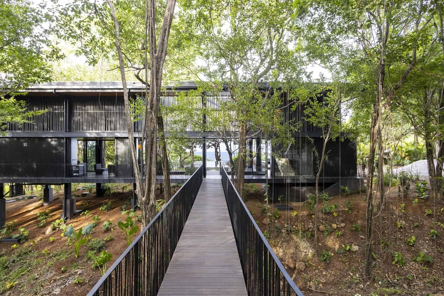 Contemporary black treehouse with modern design nestled among lush green trees and surrounded by nature, featuring a wooden walkway leading to the entrance.