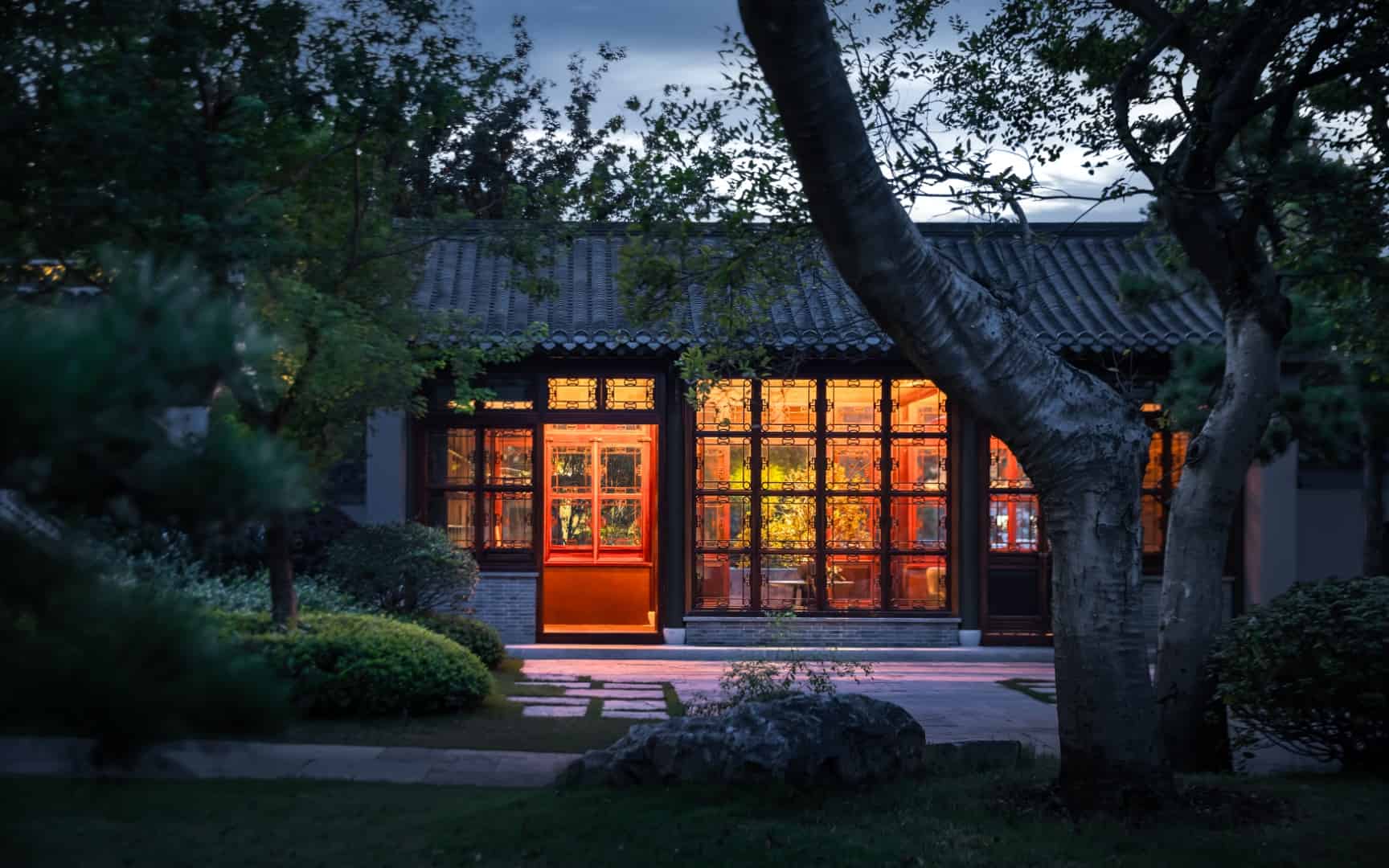 Elegant traditional Asian-style house with warm illuminated interior, surrounded by lush garden and trees at dusk, showcasing harmony between architecture and nature.