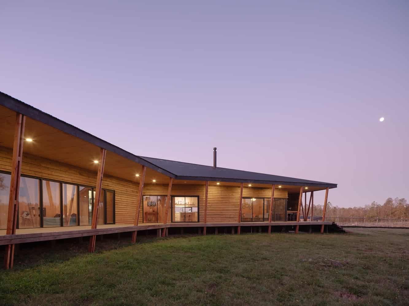 Modern wooden house with large glass windows and sloped roof, illuminated by exterior lighting at dusk, set in natural landscape, contemporary architecture design, eco-friendly building style.
