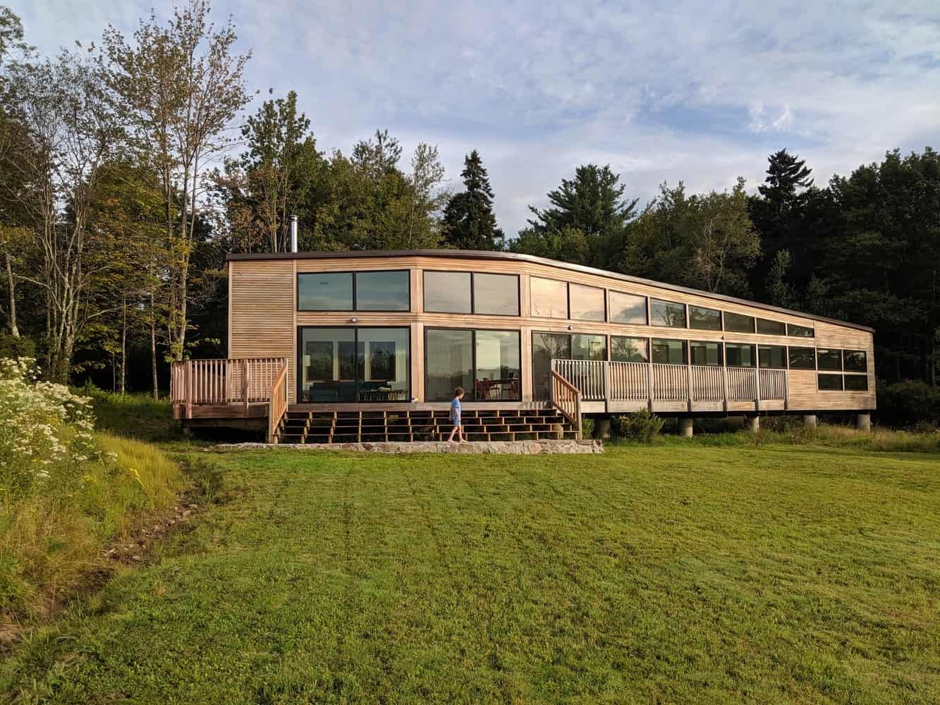 Modern wooden house with large glass windows and surrounding deck on grassy terrain, nestled among trees, showcasing contemporary architecture and eco-friendly design elements.