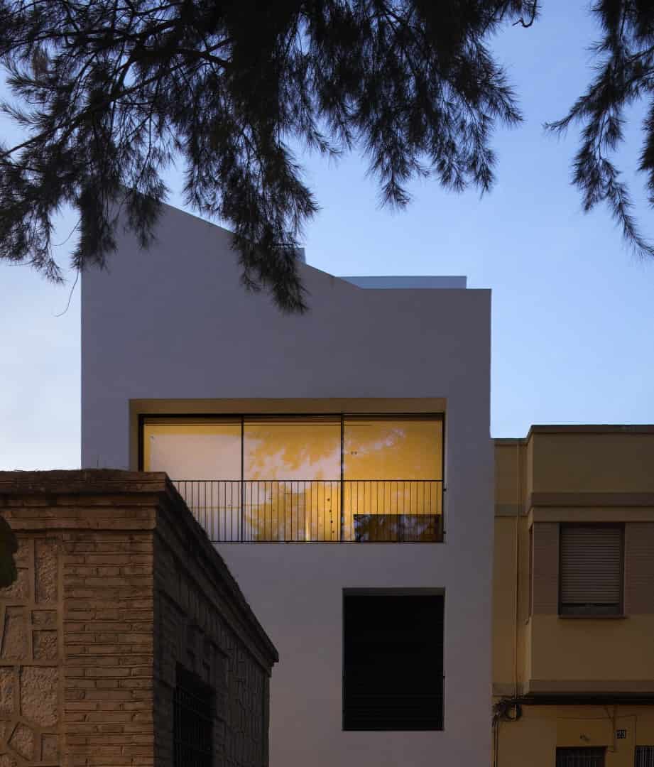 Modern minimalist white residential building with large glass windows and black metal balcony, surrounded by trees, showcasing contemporary architecture and urban design.