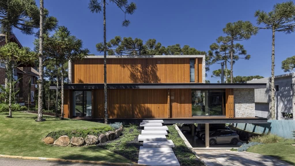Modern minimalist house with wood and concrete exterior, surrounded by tall pine trees, featuring large glass windows, a landscaped yard, and an elevated parking area.